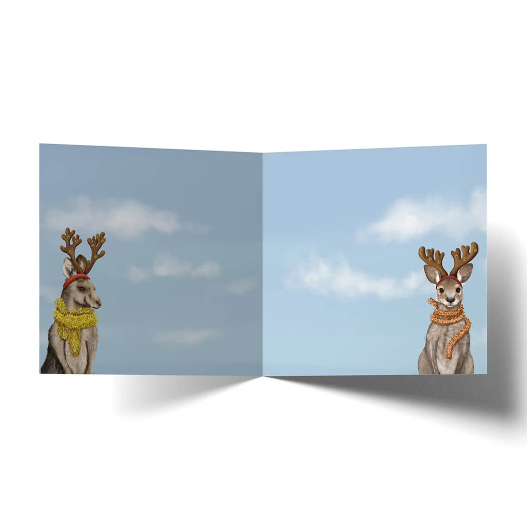 An open greeting card or art print showing a pale blue sky with clouds across both panels, with two festive kangaroos wearing antler headbands and christmas decorartions