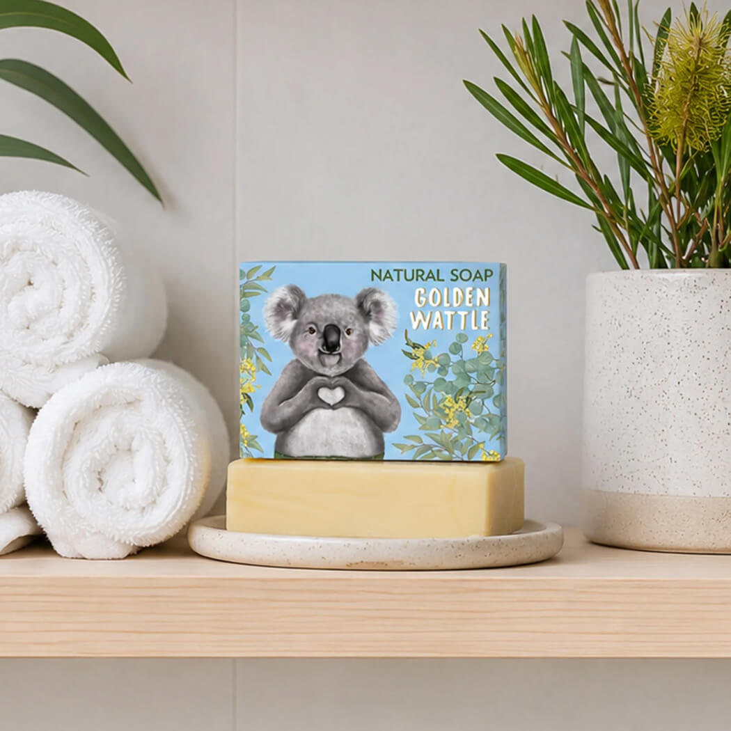 A bar of natural soap in a square box with a koala illustration and the words 'NATURAL SOAP GOLDEN WATTLE' sits on a light wood shelf beside rolled white towels, a ceramic planter, and green leaves.