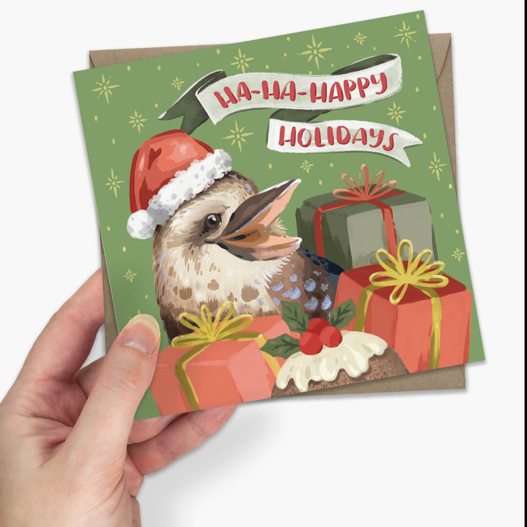 a square Christmas greeting card featuring a kookaburra wearing a Santa hat with presents and the text 'HA-HA-HAPPY HOLIDAYS' on a light green background.