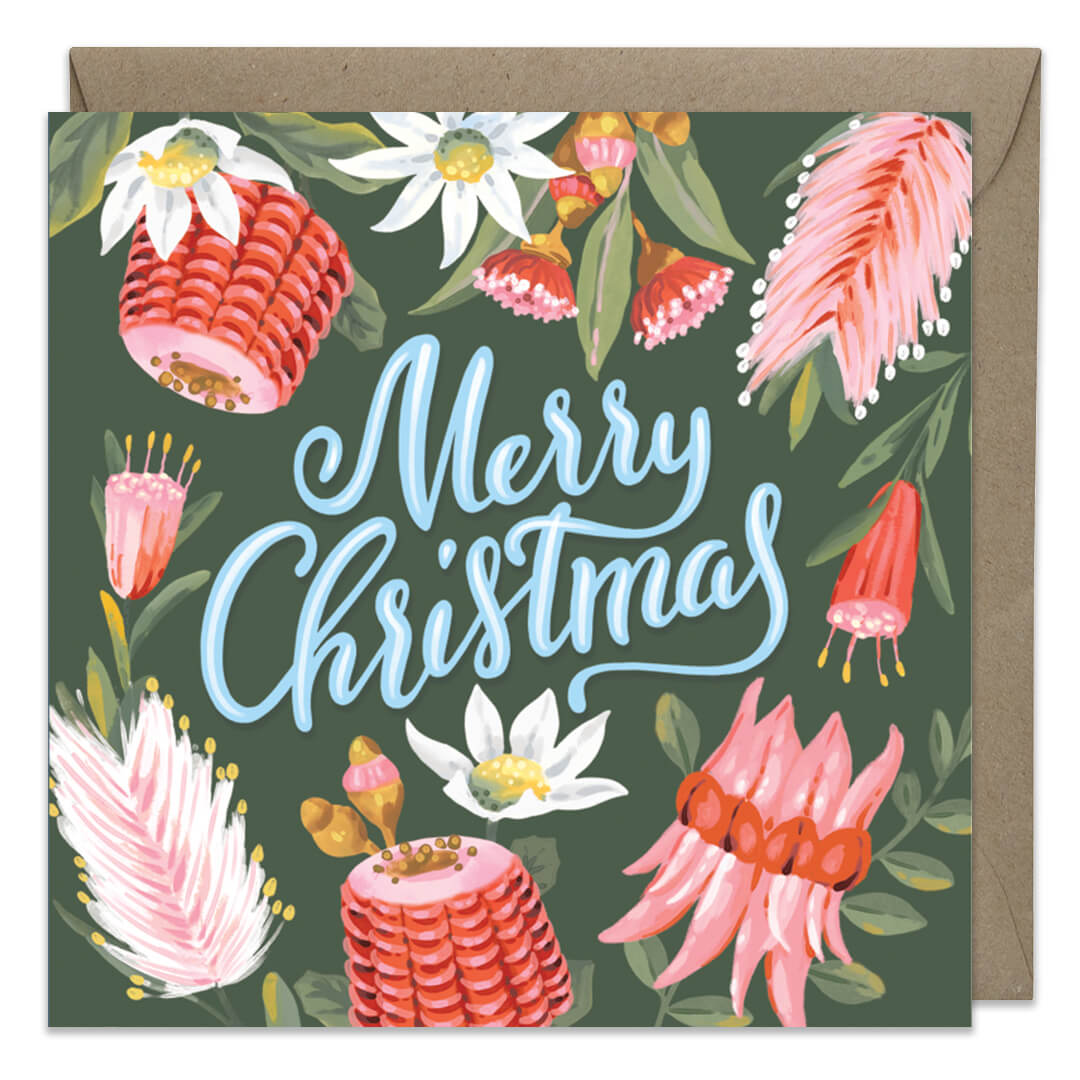 A square Christmas greeting card with a kraft envelope behind it. The card features a dark green background decorated with pink and white flowers, green leaves, and the words “Merry Christmas” in large light blue script.