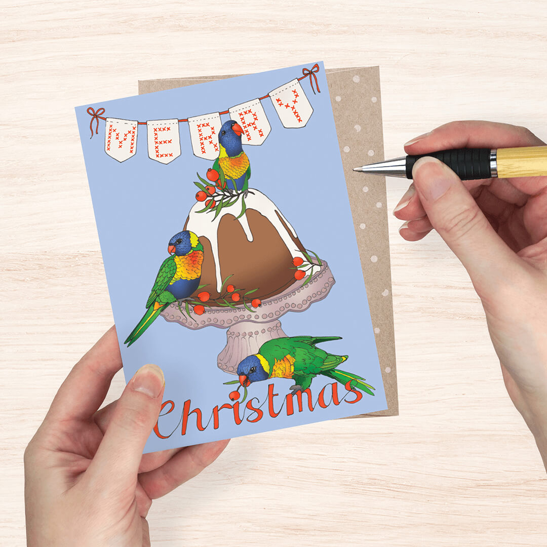 Christmas greeting card featuring a blue background, a decorated Christmas pudding, and three colorful lorikeets, with the word 'Merry Christmas' in red script. A brown envelope is behind the card, and a hand holds a pen beside it on a light wood surface.