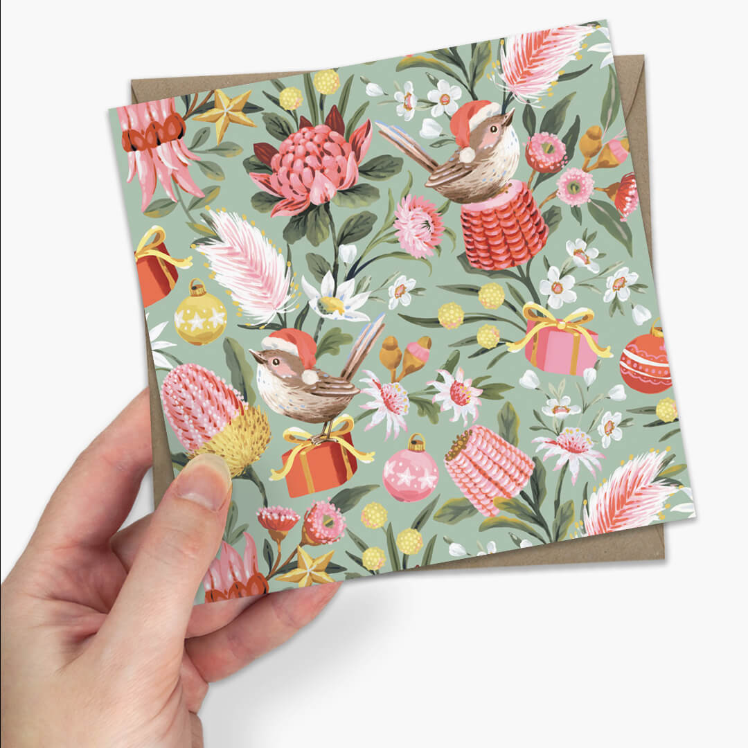 a square greeting card with a festive illustrated pattern of birds, flowers, and ornaments, with a brown envelope partially visible behind it on a light gray background.