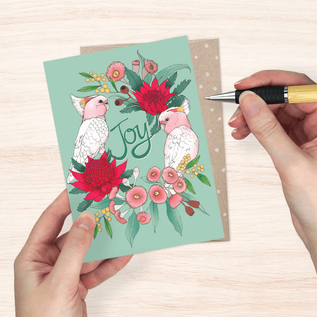 greeting card featuring two pink cockatoos, red warblers, and pink flowers with the word 'Joy' in script.