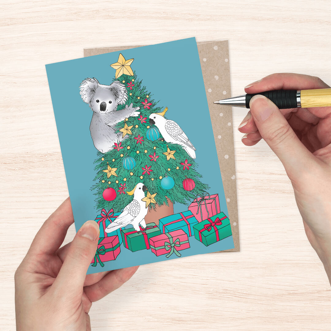 blue greeting card featuring a Christmas tree with a koala, star topper, colorful ornaments, and white cockatoos among wrapped presents. 