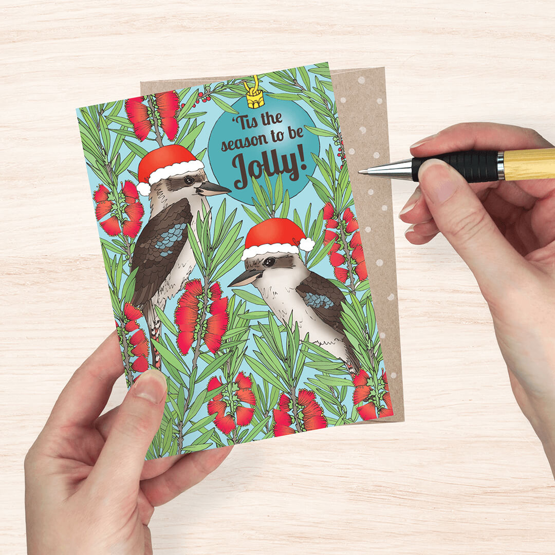 a Christmas greeting card featuring two kookaburra birds wearing Santa hats among red flowers on a light blue background, with the text 'Tie the season to be Jolly!'. A brown envelope is visible behind the card, and a hand holds a pen near it on a light wood surface.