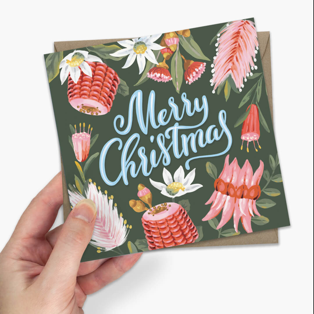 Christmas greeting card featuring illustrated pink and white flowers on a dark green background with the words “Merry Christmas” in light blue script. A brown envelope is partially visible behind the card.