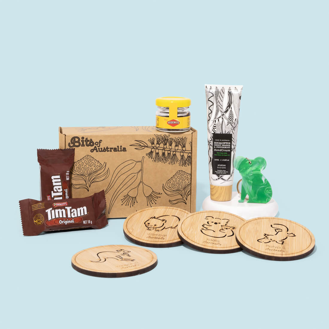A curated Australian Made gift set on a light blue background featuring a brown cardboard box labeled Bits of Australia, two TimTam candy bars, a small glass jar with a yellow lid, a white tube with black botanical illustrations, a green frog-shaped object on a white stand, and four round wooden coasters engraved with animal illustrations.