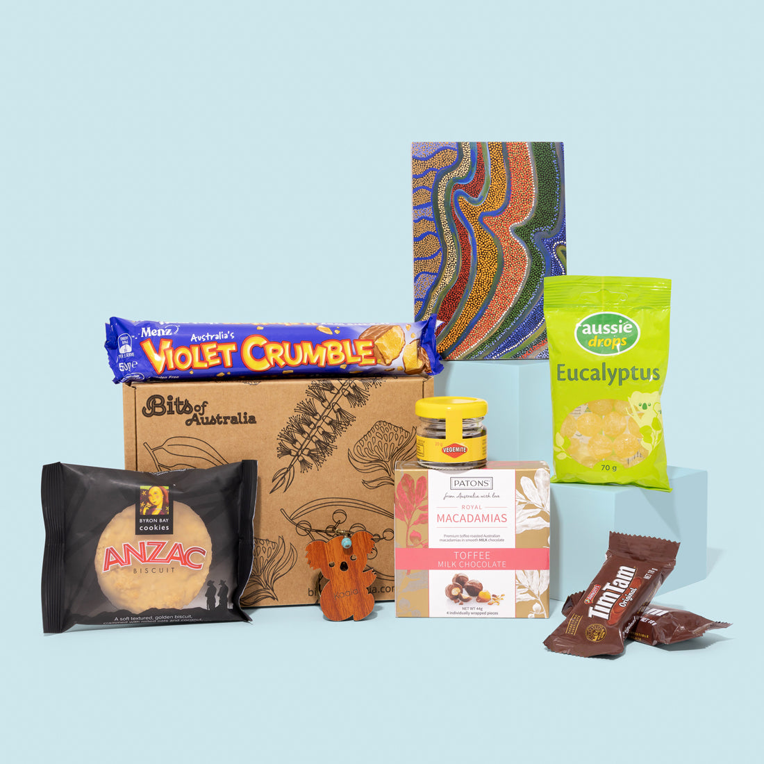 A curated gift box set featuring various packaged Australian snacks and treats. Australian gift box with Vegemite, Tim Tams, chocolate, koala keyring, eucalyptus lollies and Aboriginal art notepad